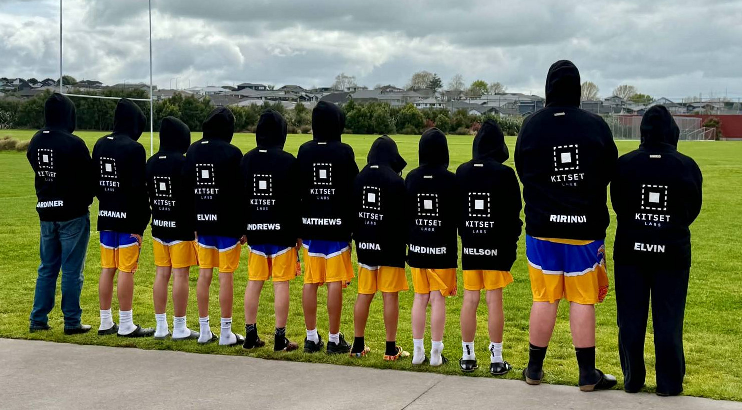 Eleven Tauranga City Basketball under 14 players standing in a row with their backs to the camera, wearing black Kitset Labs hoodies